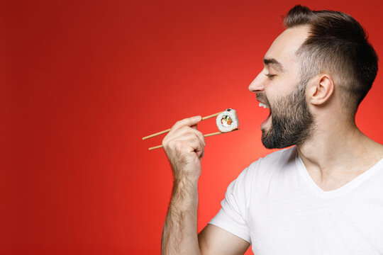 Funny Young Bearded Man 20s Wearing Casual White T-shirt Eating Hold In Hand Sticks Chopsticks With Makizushi Sushi Roll Traditional Japanese Food Isolated On Red Color Background Studio Portrait.