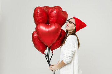 Obraz na płótnie Canvas Side view of funny young woman in sweater red hat glasses looking aside celebrating birthday holiday party hold bunch heart air inflated helium balloons isolated on white background studio portrait.