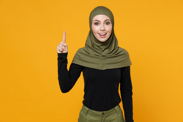 Excited young arabian muslim woman in hijab black green clothes holding index finger up with great new idea isolated on yellow color background studio portrait. People religious lifestyle concept.