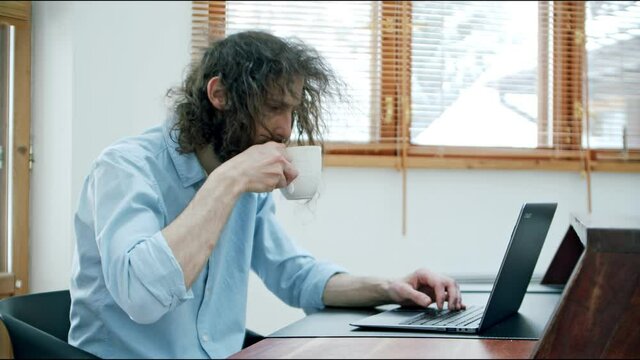 shaggy and sleepy man at the desk with a laptop drinks from a cup of coffee and becomes workable and cheerful. the drink gives a wake-up effect.