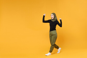 Fototapeta na wymiar Full length of happy young arabian muslim woman in hijab black green clothes doing winner gesture celebrating say yes isolated on yellow background studio portrait. People religious lifestyle concept.