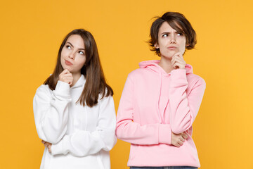 Pensive puzzled two young women friends 20s wearing casual white pink hoodies standing put hand prop up on chin looking aside up isolated on bright yellow color wall background studio portrait.