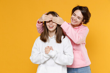 Cheerful funny surprised two young women friends 20s wearing casual basic white pink hoodies close eyes with hands, play guess who or hide and seek isolated on yellow color background studio portrait.