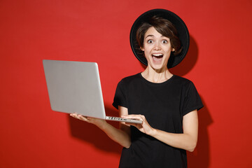 Surprised shocked amazed young brunette woman 20s years old wearing casual basic black t-shirt hat stand working on laptop pc computer say wow isolated on bright red color background studio portrait.