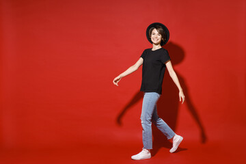 Fototapeta na wymiar Full length side view of smiling beautiful young brunette woman 20s years old wearing casual basic black t-shirt hat standing looking camera isolated on bright red color background studio portrait.