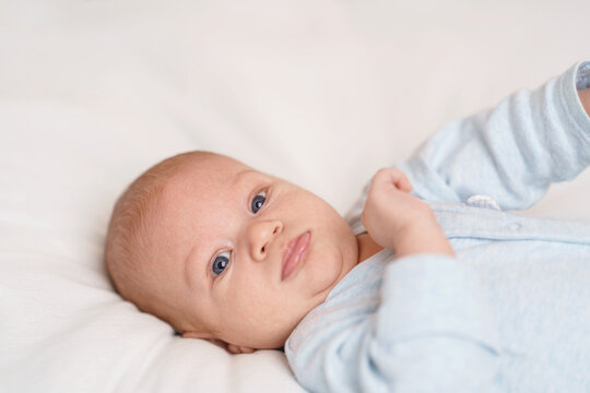 cute 2-month-old baby in blue bodysuit lies on bed and looks into frame