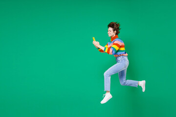 Fototapeta na wymiar Full length side view of smiling funny cheerful young brunette woman 20s in colorful sweater jumping using mobile cell phone typing sms message isolated on green color background studio portrait.