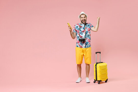 Full length of joyful young traveler tourist man using mobile phone booking hotel taxi doing winner gesture isolated on pink background. Passenger traveling on weekends. Air flight journey concept.
