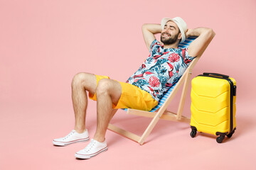 Full length of smiling young traveler tourist man in summer clothes hat sit on deck chair hold hands behind head isolated on pink background. Passenger travel on weekend. Air flight journey concept. - obrazy, fototapety, plakaty