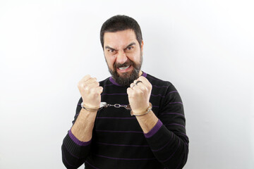 Portrait of an angry bearded man wearing casual clothes with handcuffs on white background