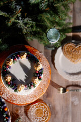 Christmas white orange mousse cake decorated cranberries, blackberries and green thuja branches on cake stand with fir tree, cookies and xmas decoration above on wooden table. Dark rustic style.