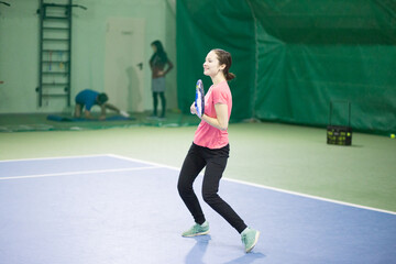 Obraz premium A young white brunette girl with a racket in her hands and in black sweatpants and a pink T-shirt is playing tennis and smiling. It is located in the indoor table tennis hall.