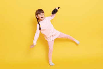 Fototapeta na wymiar Happy smiling little girl jumping and singing in microphone, small vocalist wearing casual clothing posing isolated over yellow background, child with pigtails performing.