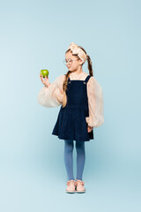 full length of little girl in dress and glasses looking at green apple on blue