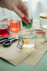 Frau gießt flüssige Ostereier Kaltfarbe Gelb in Glas mit Essig Wasser auf Pastell Papier Unterlage neben Orange, Grün, Blau, Rot in Verpackung und Schere zum Färben zu Ostern