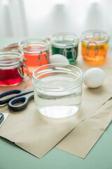 Bunte Ostereier Kaltfarben in Rot, Orange, Gelb, Blau, Grün in Verpackung auf Pastell Papier Unterlage mit Eier, Essig, Wasser, Gläser und Schere zum Färben zu Ostern
