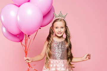 cheerful little girl in dress and crown holding balloons isolated on pink