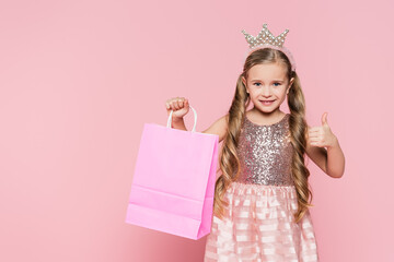 cheerful little girl in dress and crown holding shopping bag and showing thumb up isolated on pink