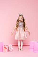Obraz na płótnie Canvas full length of shocked little girl in crown standing near presents and shopping bags on pink