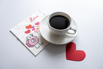 A white cup of coffee stands on a saucer and on a napkin on a white background. Also next to it is a red heart, a valentine.