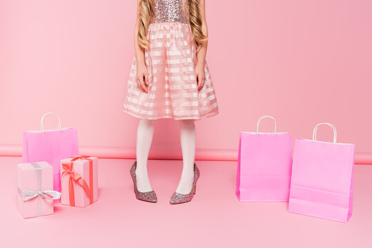 cropped view of little girl in dress standing on heels near presents and shopping bags on pink