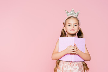smiling little girl in crown holding book while looking at camera isolated on pink