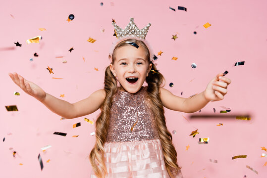 amazed little girl in crown with outstretched hands near falling confetti on pink