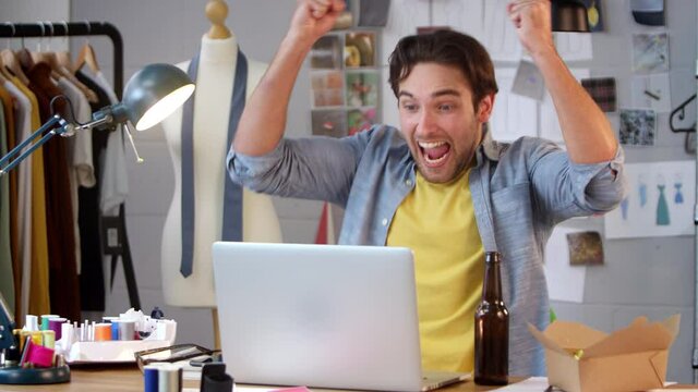 Male owner of fashion business with bottle of beer and takeaway meal watching sport on laptop in studio and celebrating goal - shot in slow motion