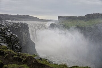 view of Dettifoss waterfall, small figures of tourists can be seen on the edge of the waterfall, cloudy weather, a cloud of water spray rises above the waterfall, nature of Iceland