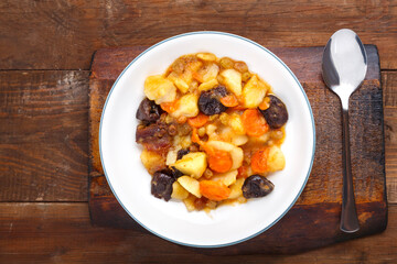 A dish of Jewish cuisine tsimes with carrots and dates in a white plate on a wooden board next to a spoon.