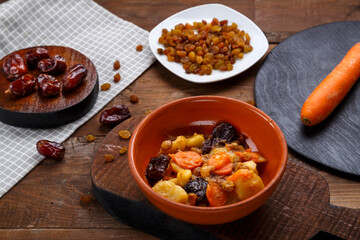 A dish of Jewish cuisine sweet tsimes with carrots with dates in a plate next to a spoon and carrots with dried fruits.