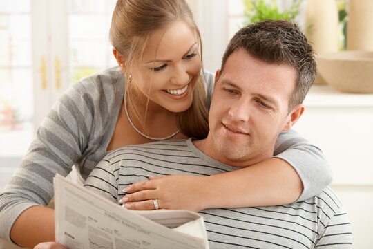 Handsome man reading newspaper at home, smiling woman hugging man looking down.