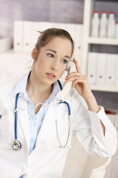 Portrait of young female doctor sitting at desk in doctors room calling, looking away, smiling.