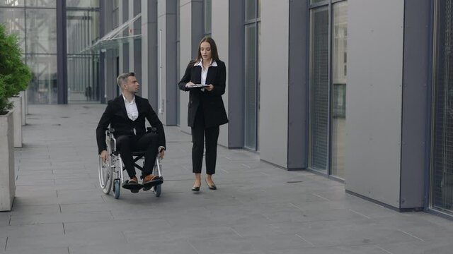Disabled male ceo executive on wheelchair and woman secretary working with documents near office building. Coworkers having conversation while discussing project and going at city street.