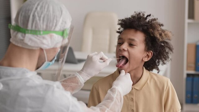 Over shoulder chest-up of teenage African boy opening his mouth for unrecognizable medical worker collecting sample of his saliva with cotton swab for analysis. Patient smiling and nodding