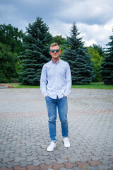 Fototapeta premium A handsome young man of European appearance wearing sunglasses is dressed in a shirt and jeans. The guy walks down the street, he is stylishly dressed