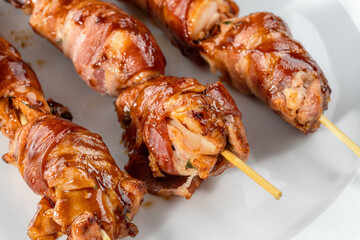 Grilled bacon-wrapped chicken tenders are laid out on a white plate. Strung on wooden skewers.