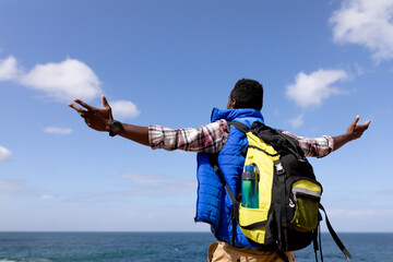 Fit afrcan american man wearing backpack hiking spreading arms on the coast