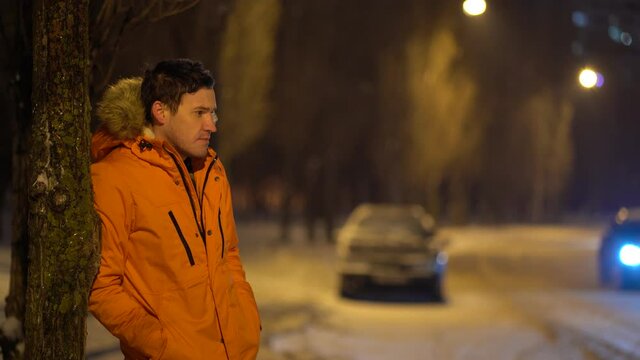 Portrait of frozen young man in orange jacket standing near tree in winter at night. Adult male looks around, standing near road in waiting for someone.