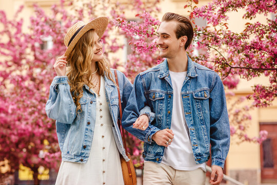 Young Happy Smiling Fashionable Couple Walking In Street Of European City, Near Pink Spring Blossom Trees. Model Wears Trendy Blue Denim Jackets