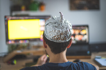 Conspiracy theory concept: young man is wearing aluminum head, sitting in front of the pc watching...