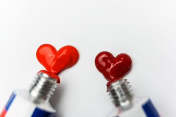 Small aluminum open tubes on a white sheet of paper with two shades of red acrylic paint, squeeze out paint in the shape of hearts, close-up view, love concept