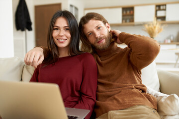 Electronic gadgets, devices and relationships concept. Stylish young bearded man embracing his beautiful wife while surfing internet together, seated on sofa in living room, smiling at camera