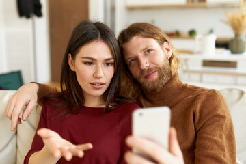 Indoor shot of stylish guy with thick beard relaxing on shoulder of his beautiful girlfriend who is sitting on sofa, holding cell phone, gesturing emotionally, having online conversation on video chat