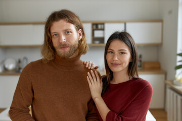 Family portrait of happy young married couple spending leisure time indoors in quarantine, bonding, standing next to each other, looking at camera with smile. Romance, love and domestic life