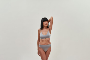 Natural beauty. Attractive half naked caucasian senior woman keeping one arm raised and looking at camera while posing in lingerie against grey background