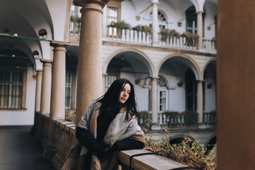 A young caucasian black-haired girl in a dark autumn coat and a wide scarf-wrap stands on the balcony with a gray colonnade and looks down thoughtfully. Lviv, Italian courtyard, Ukraine.