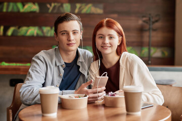 Happy teenage couple smiling at camera while listening to music together using smartphone and same pair of earphones, sitting in a cafe on a daytime
