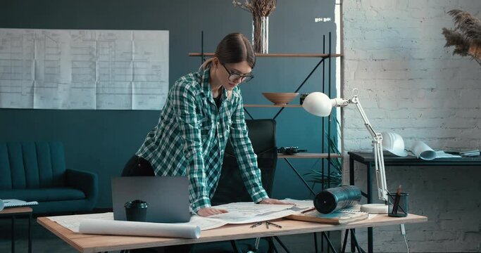 Young Designer of Interior Standing near the Workplace and Check her Project. Looking at details. Wearing a casual Shirt and Trendy eyeglasses. Beautiful Equipped Place.