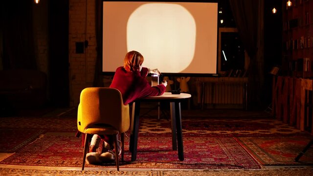 A woman is sitting on armchair and showing images on the screen with the help of slide projector standing on the table. Back view of a lady watching pictures using old fashioned cinematographic device